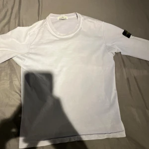Vit långärmad Stone Island tröja - Vit långärmad tröja från Stone Island med klassisk rund hals och ikonisk patch på vänster ärm. Tröjan är i mjuk bomull och har en clean, stilren look som passar perfekt till jeans. Snygg passform och diskret branding för dig som gillar streetwear.