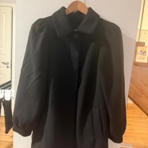 Svart kappa med puffärm - Snygg svart kappa med klassisk krage och puffade ärmar. Kappan har en enkel knäppning framtill och två fickor på sidorna. Perfekt för dig som gillar stilrena och tidlösa plagg med en twist på ärmarna. Från monki 