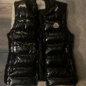Svart dunväst från Moncler - Snygg svart dunväst från Moncler med glansig finish och klassisk logga på bröstet. Västen har hög krage, två dragkedjefickor och quiltad design. Perfekt för lager-på-lager och streetstyle. Innerfoder med Monclers ikoniska serietidningsetikett.