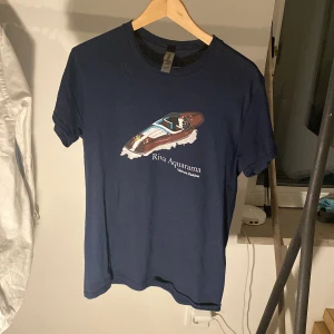 Marinblå Riva Aquarama t-shirt - Snygg marinblå t-shirt från Gildan med tryck av en klassisk Riva Aquarama-båt och texten 'Velvura Fashion' på bröstet. Gjord i heavy cotton för en skön och stadig känsla. Perfekt för dig som gillar båtliv och stilrena motiv. 199 kr