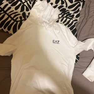 Vit hoodie från Emporio Armani EA7 - Säljer en stilren vit hoodie från Emporio Armani EA7 med logga på bröstet. Tröjan har huva med snörning och en stor magficka. Perfekt för dig som gillar en clean och modern look.