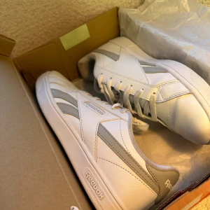 Reebok Court Retro vita sneakers storlek 38 - Snygga Reebok Court Retro sneakers i vitt och grått med klassisk design. Skorna har snörning, rund tå och platt sula. Tillverkade i syntetmaterial och skinn, perfekta för dig som gillar en clean och sportig look.