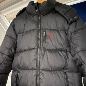 Svart pufferjacka Polo Ralph Lauren - Svart pufferjacka från Polo Ralph Lauren med röd broderad logga på bröstet. Jackan har huva, flera dragkedjor och en klassisk look. Perfekt för kalla dagar och riktigt snygg streetstil. Använd 3 gånger vilket gör den till helt ny skick! Pris kan diskuteras vid snabb affär!✅