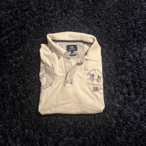 Vit piké från La Martina med brodyr - Vit pikétröja från La Martina med broderade detaljer, stora tryck och patchar på både bröst och ärm. Klassisk krage, knappar framtill och coola kontrastdetaljer. Perfekt för dig som gillar sportig och preppy stil.