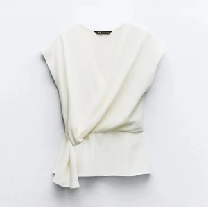 Vit omlottopp med knyt från Zara - Säljer en stilren vit omlottopp från Zara i storlek S. Toppen har en snygg omlottdesign med knyt i sidan och är ärmlös. Materialet är mjukt och har ett lätt fall, perfekt för en clean och chic look. Passformen är normal och toppen har en v-ringad front. 