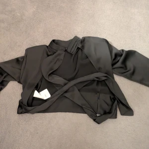 Svart omlott kavaj i silke - Snygg svart omlott blus i silkeslent material. Tröjan har lång ärm, hög krage och ett bälte som knyts i midjan.