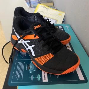 Svarta och orangea Asics sneakers - Säljer ett par sportiga Asics sneakers i svart med orangea och vita detaljer. Skorna har coolt hexagonmönster på ovandelen, snörning och platt sula. Perfekta för dig som gillar att sticka ut med färg och vill ha bekväma skor till träning eller vardag.