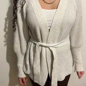 Vit stickad kofta med bälte - Mysig vit stickad kofta med lång ärm och bred ribbad struktur. Koftan har ett matchande bälte i midjan som kan knytas för extra stilpoäng. Perfekt att slänga på sig när det är lite kyligt ute. Snygg v-ringad öppning framtill och skön passform.