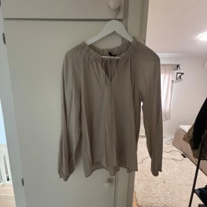 Beige blus med knyt från Vero Moda - Säljer en beige blus från Vero Moda med långa ärmar och snygg rynkad halsringning med knyt framtill. Blusen har en luftig passform och är perfekt för dig som gillar en enkel men stilren look. Bra skick