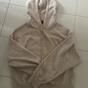 Beige stickad hoodie med huva - Mysig beige stickad hoodie med huva och snörning. Tröjan har långa ärmar och ribbade muddar vid ärmslut och nederkant. Perfekt för kyliga dagar och enkel att matcha med jeans eller mjukisbyxor.