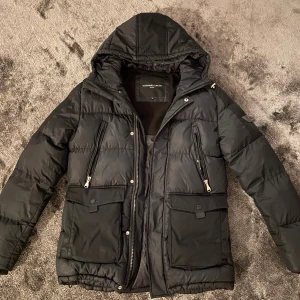 Svart pufferjacka Alessandro Zavetti - Svart pufferjacka från Alessandro Zavetti med huva och stora fickor framtill. Jackan har dragkedjor på bröstfickorna och tryckknappar på de nedre fickorna. Insidan har ett snyggt mönster med logga. Perfekt för kalla dagar och riktigt stilren.