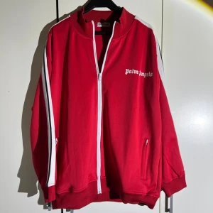 Röd Palm Angels track jacket - Palm Angels track jacket i rött med vita och svarta ränder längs ärmarna. Jackan har dragkedja framtill, ståkrage och fickor med dragkedja. Loggan är tryckt på bröstet. Perfekt för en sportig och streetwear-inspirerad look. Storlek L men passar även M. Fick den i julklapp men aldrig använt då den var för stor. 