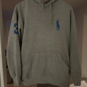 Grå hoodie från Ralph Lauren - Säljer en stilren grå hoodie från Ralph Lauren med blå logga på bröstet och stort nummer 3 på ärmen och ryggen. Tröjan är bra skick förutom att den är lite sliten på smörerna längst ut annars i bra skick!