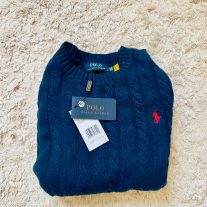 Mörkblå stickad tröja Polo Ralph Lauren - Kabelstickad mörkblå tröja från Polo Ralph Lauren med klassisk röd broderad logga på bröstet. Tröjan har rund halsringning och ribbade muddar. Perfekt för dig som gillar en tidlös och clean stil.