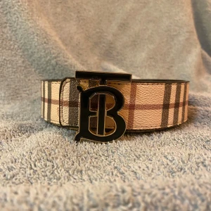 Burberry bälte med rutigt mönster - Säljer ett Burberry bälte med klassiskt rutigt mönster i beige, brun och svart. Bältet har en bred form och en stor metallspänne med Burberrys ikoniska TB-logga i svart och guld. Materialet är skinn med tryckt mönster och exklusiv känsla.
