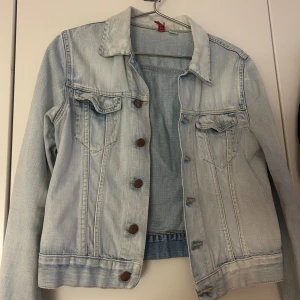 Ljusblå jeansjacka med knappar - Snygg ljusblå jeansjacka från H&M med klassisk krage och bröstfickor med lock. Jackan har bruna knappar framtill och är långärmad. Perfekt att slänga över en hoodie eller t-shirt för en chill look. 