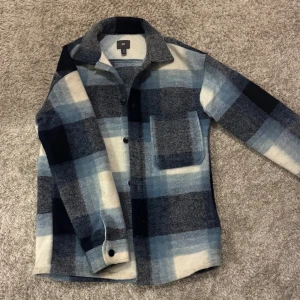 Rutig overshirt från H&M i blått - Säljer en snygg rutig overshirt från H&M i olika nyanser av blått, grått och vitt. Skjortan har en stor bröstficka, klassisk krage och knappar framtill. Materialet är mjukt och tjockt, perfekt för lager-på-lager. Passar dig som gillar streetwear och chill stil.