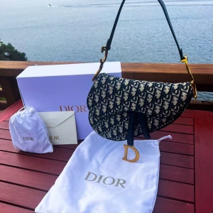 Dior Saddle axelväska med monogram - Säljer en ikonisk Dior Saddle axelväska med blå och guld monogrammönster. Väskan har guldfärgade metalldetaljer, D-hänge och justerbar axelrem i svart skinn. Snygg, kurvad form och klassisk Dior-logga över hela väskan. Kommer med dustbag och originalförpackning.