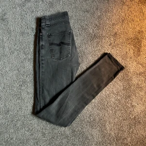 Svarta skinny jeans från Nudie Jeans - Säljer ett par svarta skinny jeans från Nudie Jeans i storlek 27/34. Jeansen har klassisk femficksdesign, svarta sömmar och Nudie-loggan broderad på bakfickan. Snygg passform med smala ben och normal midja. Perfekta till sneakers eller boots.