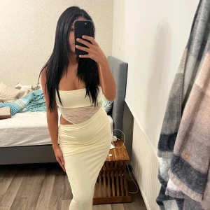 Ljus långklänning med spetsdetalj - Säljer en snygg ljus långklänning med spetsdetalj i midjan och cut-out på sidan. Klänningen är ärmlös och har en figurnära passform som framhäver siluetten. Perfekt för dig som vill sticka ut med en unik och trendig look.