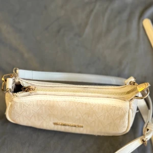Vit axelväska från Michael Kors - Snygg vit axelväska från Michael Kors med justerbar rem och guldfärgade detaljer. Väskan har ett diskret mönster med MK-loggor och dragkedja upptill. Perfekt storlek för det viktigaste och passar till många olika outfits.