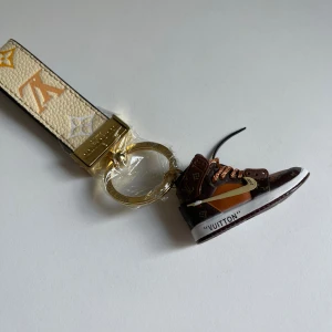  Vuitton nyckelring med sneaker - Cool  Vuitton nyckelring med beige och guldigt band i skinn och en miniatyr sneaker i brunt, orange och vitt med monogram. Snygg accessoar till väskan eller nycklarna, med guldfärgad metallring och detaljer.