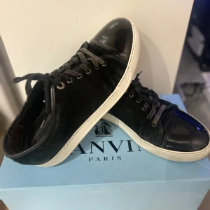   Lanvin svarta  - Snygga svarta sneakers från Lanvin Paris med vit platt sula och snörning. Skorna har en blank tå i skinn och resten av ovandelen är i mocka, vilket ger en cool kontrast. Perfekta för dig som gillar stilrena och lyxiga detaljer.                                              Bara att skriva vid frågor och funderingar.  Priset är ej hugget i sten 