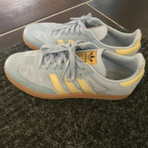 Adidas Samba ljusblå/gula sneakers - Säljer ett par Adidas Samba sneakers i ljusblått med gula detaljer och klassiska tre ränder på sidan. Skorna har snörning, platt gummisula och är tillverkade i en mix av syntet och skinn. Perfekta för dig som gillar retrostil och vill sticka ut med färg. Använda fåtal gånger❤️❤️köpta för 950