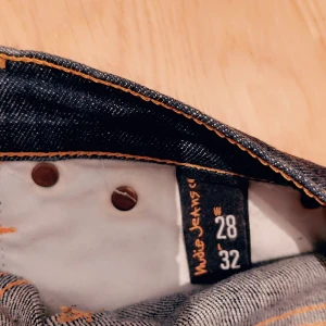 Mörkblå  jeans från Nudie Jeans - Säljer ett par mörkblå skinny jeans från Nudie Jeans med klassiska orange sömmar och snygga bakfickor med vågig brodyr. Jeansen har fem fickor och är tillverkade i slitstarkt denimtyg. Perfekt passform för dig som gillar mer slimfit.. Modellen på bilden är 172 och väger 62 kg. Sitter tight i midjan och på benen. 