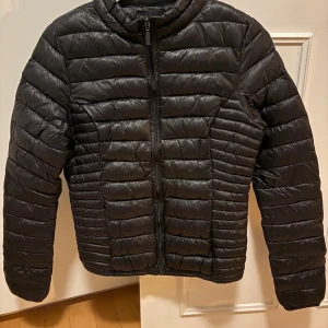 Svart lätt dunjacka från Neo Noir XS - Säljer en svart lätt dunjacka från Neo Noir i storlek XS. Jackan har horisontella quiltade sömmar, dragkedja framtill och en klassisk krage. Perfekt för kyliga dagar och riktigt lätt att matcha med allt.