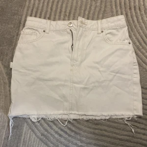 Vit jeanskjol från Bershka - Vit denimkjol från Bershka med rå kant nertill och klassiska jeansdetaljer som fickor fram och bak samt bälteshällor. Kjolen har dragkedja och knapp framtill och en cool, avslappnad look. Perfekt för dig som gillar streetstyle och enkel design.