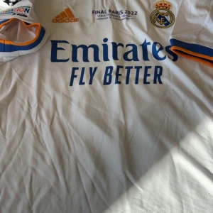 Real Madrid Kroos Final Paris 2022 tröja - Säljer en officiell Real Madrid matchtröja från Champions League-finalen 2022 med Kroos och nummer 8 på ryggen. Tröjan är vit med blå och orange detaljer, Adidas-logga och Aeroready-material som andas. Real Madrid-emblem och tryck från finalen på bröstet.