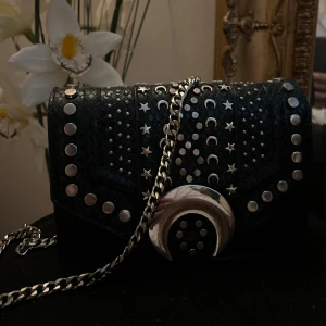 Svart axelväska med silverdetaljer - Zara - Otroligt fin svart axelväska med kedja i silver och präglad yta som liknar reptilmönster. Väskan har massor av silvernitar, stjärnor och månar på framsidan samt en stor måndetalj som spänne. Perfekt för dig som gillar edgy och unik stil. Väskan är använd ett fåtal gånger och är HELT SLUTSÅLD! Inhandlades för 450 kr, men jag säljer för billigare!💕