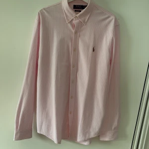 Rosa randig skjorta från Polo Ralph Lauren - Snygg rosa skjorta från Polo Ralph Lauren med vita ränder och klassisk button-down krage. Skjortan har lång ärm, broderad logga på bröstet och knappar framtill. Tillverkad i mjuk bomull, perfekt för en clean och fräsch look. Nypris 2000