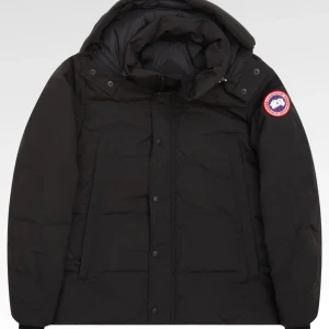 Svart dunjacka från Canada Goose - Svart dunjacka från Canada Goose med huva och klassisk logga på ärmen. Jackan har dragkedja och knappar framtill, två stora fickor och en hög krage. Perfekt för kalla vinterdagar och riktigt snygg streetstil.