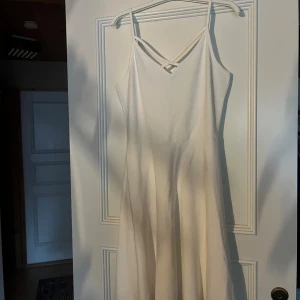 Vit klänning med smala axelband - Säljer en vit klänning med smala axelband och snyggt korsad detalj vid halsen. Klänningen har en lätt utsvängd kjol och ärmlös design som ger en fräsch och enkel look. Perfekt för varma dagar och sommarens alla tillfällen. Använd 1 gång.