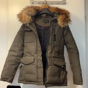 Olivgrön dunjacka  från Hollies - Jag säljer min olivgröna dunjacka från Hollies med stor huva och fluffig äktapäls. Jackan har flera fickor med dragkedja, Perfekt för kalla dagar och riktigt varm. Den är i storlek 34/XS. Den är i mycket bra skick och har använts i en vinter. Den säljs för 2500 men priset kan diskuteras vid snabb affär.