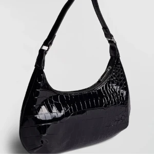 Svart handväska  - Snygg svart och enkel axelväska med glansig finish. Väskan har en böjd form och ett brett axelband med silverfärgade detaljer. 