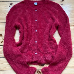 Röd fluffig kofta med volangdetaljer - Supermjuk och fluffig röd kofta med pärlemorknappar framtill. Koftan har långa ärmar och volangdetaljer vid ärmslut och nederkant som ger en extra söt touch. Perfekt för att mysa till hösten eller vintern. I storlek 158/164 från Lindex 