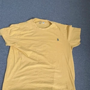 Gul t-shirt från Polo Ralph Lauren - Snygg gul t-shirt från Polo Ralph Lauren med klassisk blå broderad logga på bröstet. Modellen är kortärmad och har en rund halsringning. Tillverkad i mjuk bomull som känns skön mot huden. Perfekt för dig som gillar stilrena och ikoniska plagg.