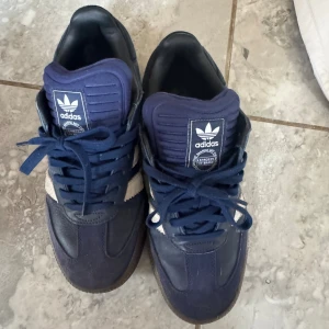 Adidas Samba sneakers marinblå - Säljer ett par klassiska Adidas Samba sneakers i marinblått med vita detaljer och snörning. Skorna har en retro vibe med rund tå och platt sula, perfekt för dig som gillar streetstyle. Materialet är en mix av skinn och syntet, och de har den ikoniska Adidas-loggan på plösen.
