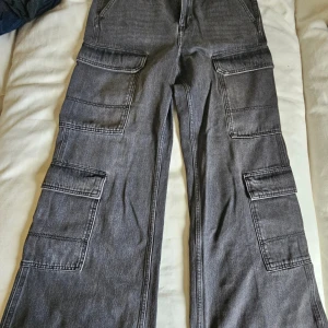 Grå cargopants från H&M Divided - Säljer ett par grå cargopants från H&M Divided med breda ben och flera stora fickor på sidorna. Byxorna har en loose passform och är tillverkade i jeansmaterial. Perfekt för dig som gillar streetstyle och vill ha mycket plats för prylar.