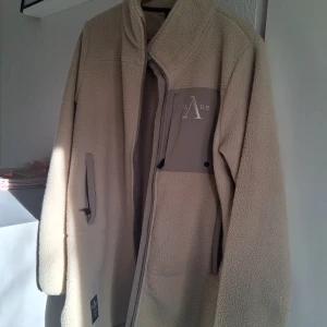 Beige teddyjacka från Slade Couture - Säljer en beige teddyjacka från Slade Couture i storlek EU 44. Jackan har hög krage, långa ärmar och praktiska fickor med dragkedja. Materialet är mjuk fleece och jackan har en cool patch med logga på bröstet och nedtill. Perfekt för dig som gillar chill och stilren vibe.