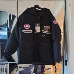Svart dunjacka från Canada Goose - Canada Goose dunjacka i svart med reflekterande detaljer och klassiska Arctic Program-patchar på bröst och ärm. Jackan har flera fickor, huva och dragkedja i sidan. Materialet är nylon och fyllningen är dun. Perfekt för kalla vinterdagar.
