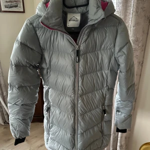 Grå McKinley pufferjacka med huva - Säljer en grå pufferjacka från McKinley med huva och rosa foder. Jackan har dragkedja framtill och vid fickorna, samt långa ärmar med svarta muddar. Perfekt för kalla dagar och har en clean, sportig look.