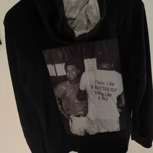 Svart hoodie med tryck från Limitato - Svart hoodie från Limitato med dragkedja framtill och två fickor med dragkedjor. Baksidan har ett stort svartvitt tryck med texten 'Float Like A BUTTER FLY Sting Like A BEE'. Mjuk insida och huva med ljusgrått foder.                                          (Går inte att få tag på längre!)