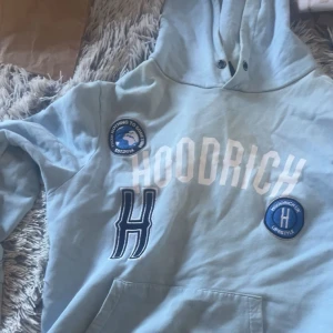 Ljusblå hoodie från Hoodrich - Säljer en ljusblå hoodie från Hoodrich med stora vita tryck och broderade patches framtill. Tröjan har huva, känguruficka och långa ärmar. Perfekt för dig som gillar streetwear och vill ha en chill look.