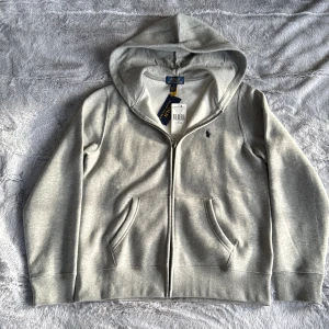 Grå hoodie från Polo Ralph Lauren - Snygg ljusgrå hoodie från Polo Ralph Lauren med dragkedja och huva. Tröjan har två fickor framtill och klassisk broderad logga på bröstet. Tillverkad i mjuk bomullsblandning, perfekt för chill dagar eller när du vill ha en clean look. Köpt på kidsbrandstor och den var för liten för mig så jag säljer den 