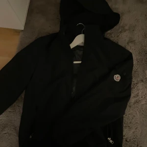Svart vindjacka från Moncler med huva - Säljer en svart vindjacka från Moncler med huva och dragkedja. Jackan har Moncler-logga på ärmen och coolt serietryck på insidan. Perfekt för dig som vill ha en stilren och funktionell jacka med streetkänsla.