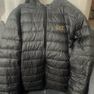  EA7 Emporio Armani svart jacka  - Svart pufferjacka från EA7 Emporio Armani med guldfärgad logga på bröstet. Jackan har huva, dragkedja framtill och quiltad design. Perfekt för kalla dagar och har en clean, sportig look.